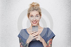 Young cute girl with tattoos over her arms wearing casual blue t-shirt hanging smartphone and smiling