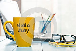 Hello July - inscription at yellow morning coffee or tea cup at business office background. Summer month, Calendar