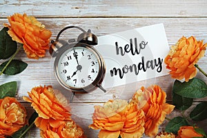 Hello Monday card and alarm clock with orange flower decoration on wooden background