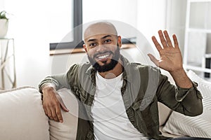 Hello. Portrait of happy mature latin man waving hand and smiling to camera, sitting on sofa at home