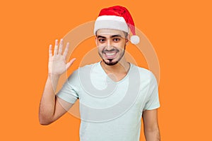 Hello! Portrait of positive handsome bearded man in santa hat and casual white t-shirt waving raised hand and saying hi to camera