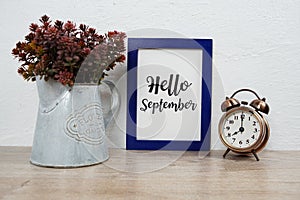 Hello September text with alarm clock and artificial plant decoration on wooden table and white wall background