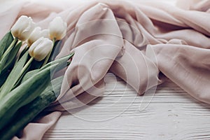 hello spring. stylish white tulips on beige soft fabric on rustic wooden table background. soft light, tenderness atmospheric mom