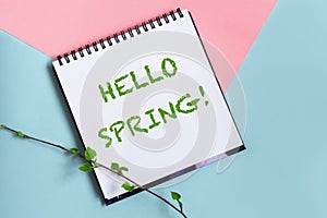 Hello spring words message on spiral scketchbook white paper on pastel pink and blue background. Twig with fresh leaves