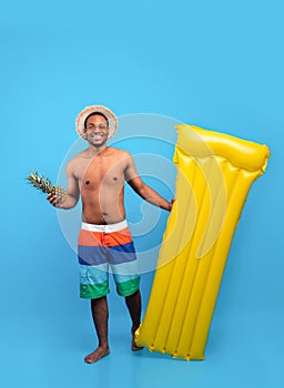 Hello summer. Full length portrait of black man in swimwear holding pineapple and inflatable lilo on blue background