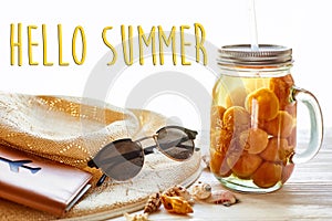 hello summer text on yellow cocktail juice with apricot, hat, sunglasses and shells on white rustic wooden background in light. s