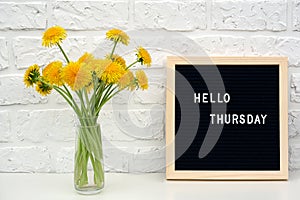 Hello Thursday words on black letter board and bouquet of yellow dandelions flowers on table against white brick wall. Concept