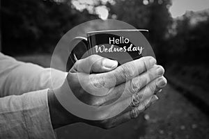 Hello Wednesday text on cup of tea or coffee in hands of person in black and white background.