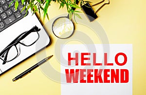 HELLO WEEKEND written on white paper near a computer, magnifier, glasses, pens on a yellow background. Motivational concept. Flat