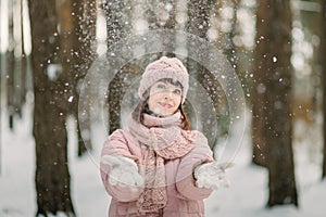 Hello winter. Smiling happy young woman in pink coat, cap and scarf, outdoors in the forest in winter, throwing up snow