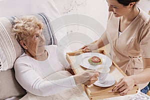 Helpful volunteer in beige uniform serving coffee to senior female patient