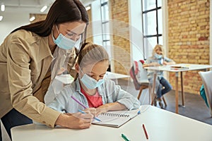 Helpful young female teacher wearing protective face mask helping little girl doing sums. Kids studying in elementary