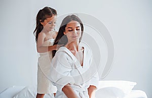 Helping to made hairstyle. Young mother with her daugher have beauty day indoors in white room