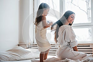 Helping to made hairstyle. Young mother with her daugher have beauty day indoors in white room