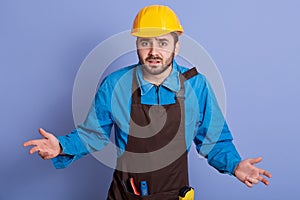 Helpless bearded engineer wearing yellow protective helmet spreading his hands to sides, looks sad and upset, does not know what