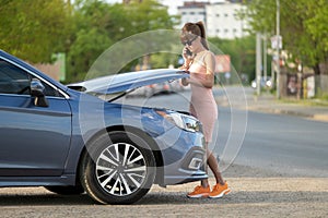 Helpless woman standing near her car with open bonnet calling road service for help. Young female driver having trouble with