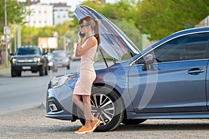 Helpless woman standing near her car with open bonnet calling road service for help. Young female driver having trouble