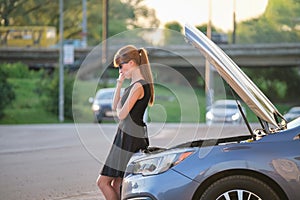 Helpless woman standing near her car with open bonnet inspecting broken motor. Young female driver having trouble with