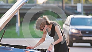 Helpless woman standing near her car with open bonnet inspecting broken motor. Young female driver having trouble with