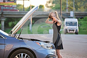 Helpless woman standing near her car with open bonnet inspecting broken motor. Young female driver having trouble with