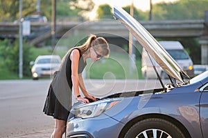 Helpless woman standing near her car with open bonnet inspecting broken motor. Young female driver having trouble with