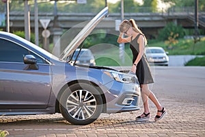 Helpless woman standing near her car with open bonnet inspecting broken motor. Young female driver having trouble with