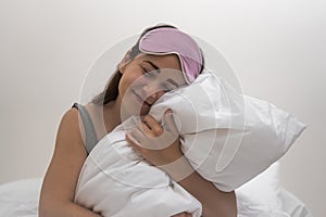 In her bedroom lady with a sleep mask smiles, cherishing a fluffy white pillow in the morning.