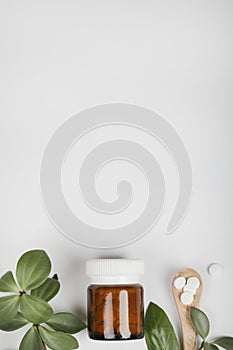 Herbal pills in glass bottle, wooden spoon and green plant leaves on white background.