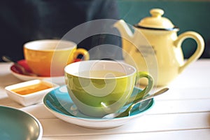 Herbal tea in green cup, honey and yellow teapot on white wooden table in cafe. A cup of hot tea with steam. Natural light