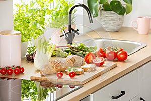 Herbs, bread and tomatoes on a cutting board next to a sink in a kitchen interior