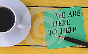 WE ARE HERE TO HELP - words on a green sheet on a yellow wooden background with a pen and a cup of coffee