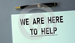 WE ARE HERE TO HELP - words on a piece of paper with a pen on a gray background photo