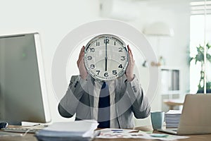 Hes got time on his mind. an unidentifiable businessman holding a clock in front of his face while sitting at his desk.