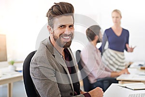 Hes one confident exec. A businessman sitting at his desk with his coworkers in the background.