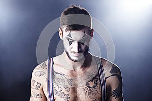 Hes one sinister-looking clown. Studio shot of a tattooed man with clown makeup on.