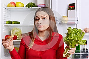 Hesitant young female model holds lettuce and sandwhich in hands, doesn`t know what to choose, can`t keep to diet, stands near ref