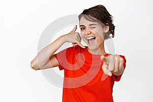 Hey call me maby. Coquettish and flirty smiling girl winking, showing mobile phone gesture and pointing finger at camera