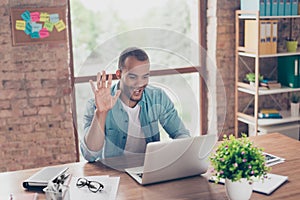 Hey there! Young cheerful afro american guy is waving in camera while having video call sitting at his work station in casual clot