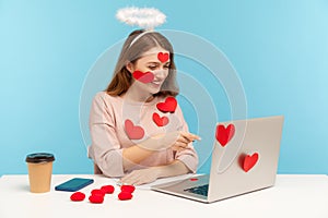 Hey you! Attractive angelic woman with kind expression sitting covered with sticker love hearts, pointing to laptop screen