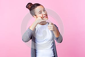 Hey you, call me! Portrait of adorable brunette teen girl smiling playfully, making telephone gesture. isolated on pink background