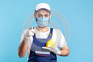 Hey you! Portrait of handyman, mover guy in workwear and gloves holding paper house and pointing to camera