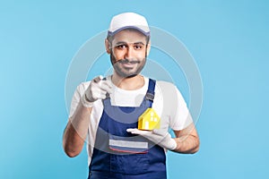 Hey you! Portrait of handyman, mover guy in workwear and gloves holding paper house and pointing to camera