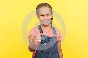 Hey you! Portrait of happy little girl in denim overalls pointing to camera and looking with positive smiling expression