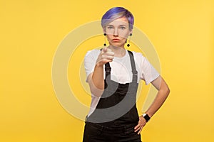 Hey you! Portrait of hipster woman pointing at camera and looking strict rigid. yellow background, studio shot