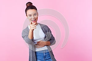 Hey you! Portrait of joyful brunette teenage girl pointing at camera and laughing out loud. studio shot, pink background