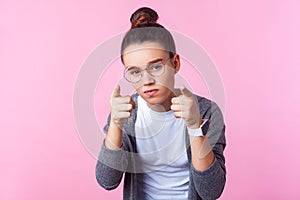 Hey you! Portrait of sophisticated brunette teenage girl in round eyeglasses pointing at camera. pink background