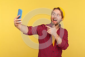 Hey you there! Friendly hipster guy in checkered shirt pointing finger to cellphone, smiling talking video call on mobile phone