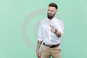 Hey you! Young adult bearded man, pointing finger and looking at camera. On light green background. Indoor