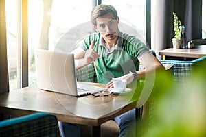 Hey you! Young serious businessman in green t-shirt sitting and working on laptop, looking at camera with warning sign