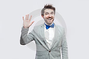 Hi, Portrait of excited happy handsome bearded man in casual grey suit and blue bow tie standing and looking at camera, greeting